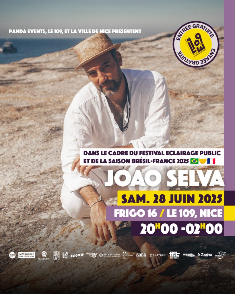 oão Selva assis sur des rochers au bord de la mer, portant un chapeau en paille et une chemise blanche, les bras croisés, sous un ciel dégagé.