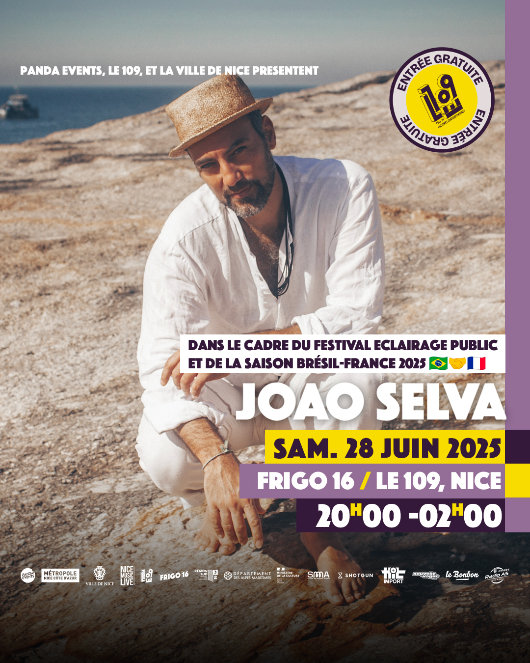 oão Selva assis sur des rochers au bord de la mer, portant un chapeau en paille et une chemise blanche, les bras croisés, sous un ciel dégagé.